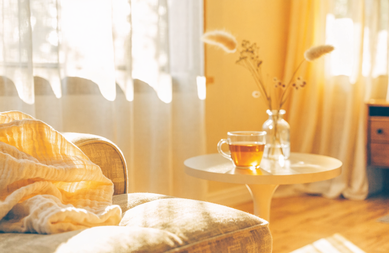 Peaceful sunlit living room in early morning with soft curtains, a folded blanket and a cup of tea