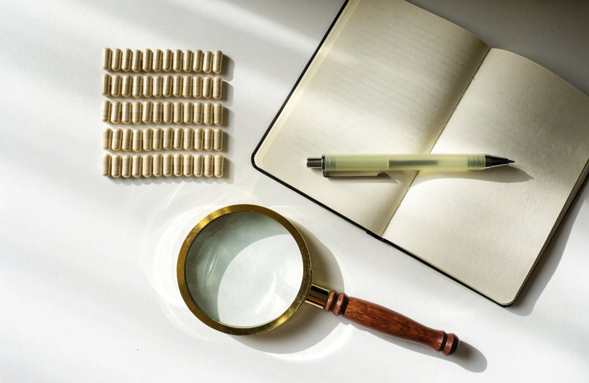 Flat lay of supplement capsules, notebook and magnifying glass on a white desk