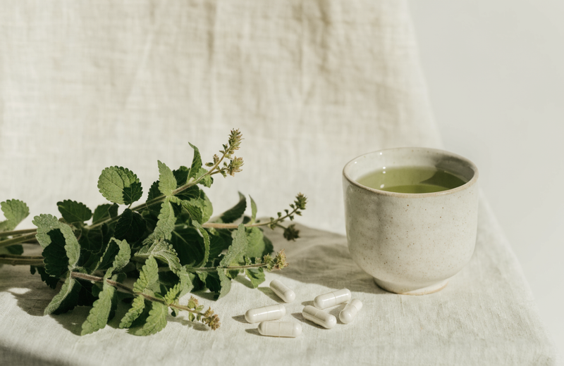 Lemon balm sprigs, supplement capsules and a cup of green tea on pale linen
