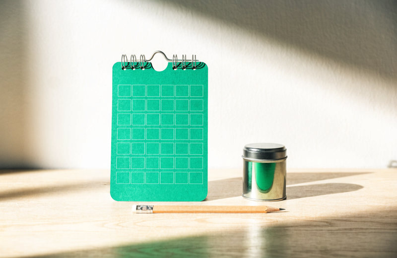 A blank two-week calendar with a supplement tin + pencil on a wooden desk in morning light