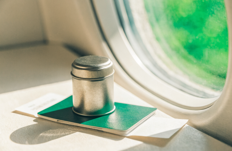 A pocket supplement tin resting on a passport + boarding pass beside an airplane window