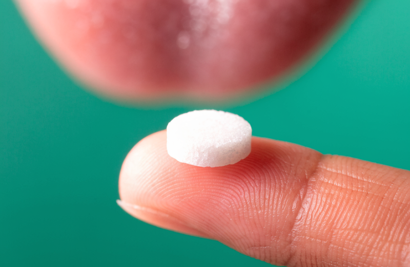 A single white sublingual mint held on a fingertip against a deep green backdrop