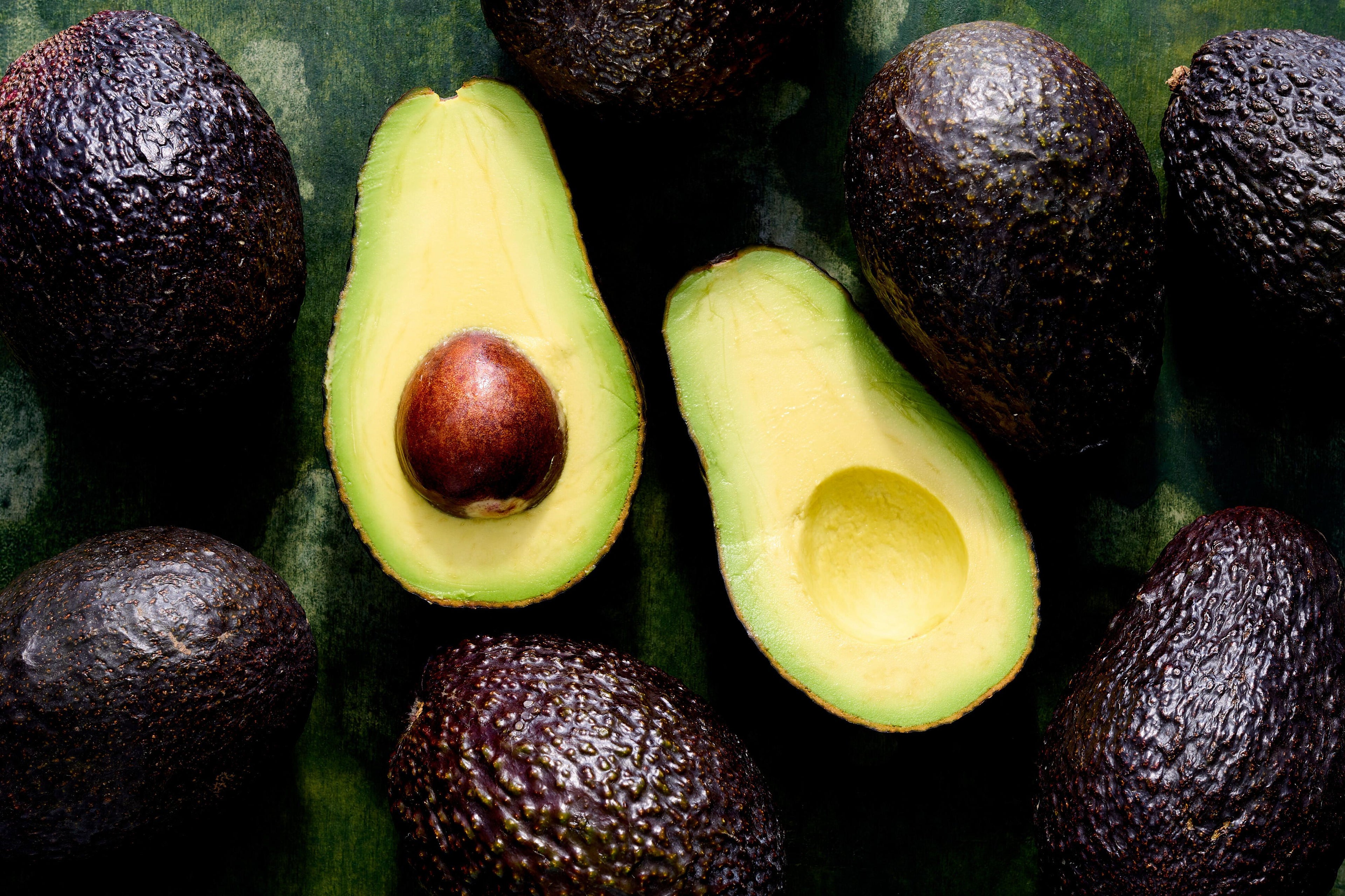 Avocado with a cut in half surrounded by whole avocados on a dark background